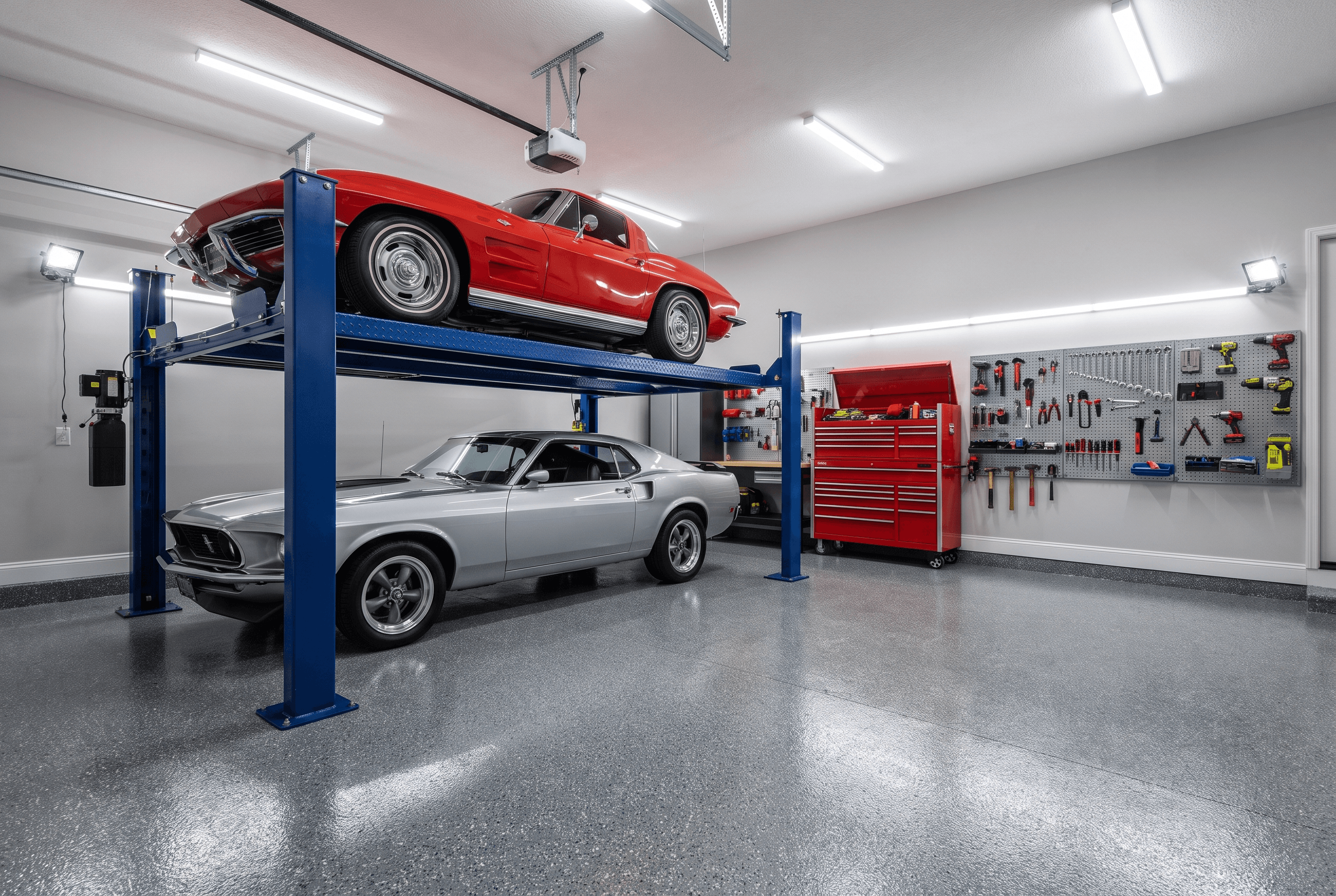A blue four-post storage lift holding a red classic sports car above a silver classic fastback in a home garage with an epoxy floor and a red tool chest